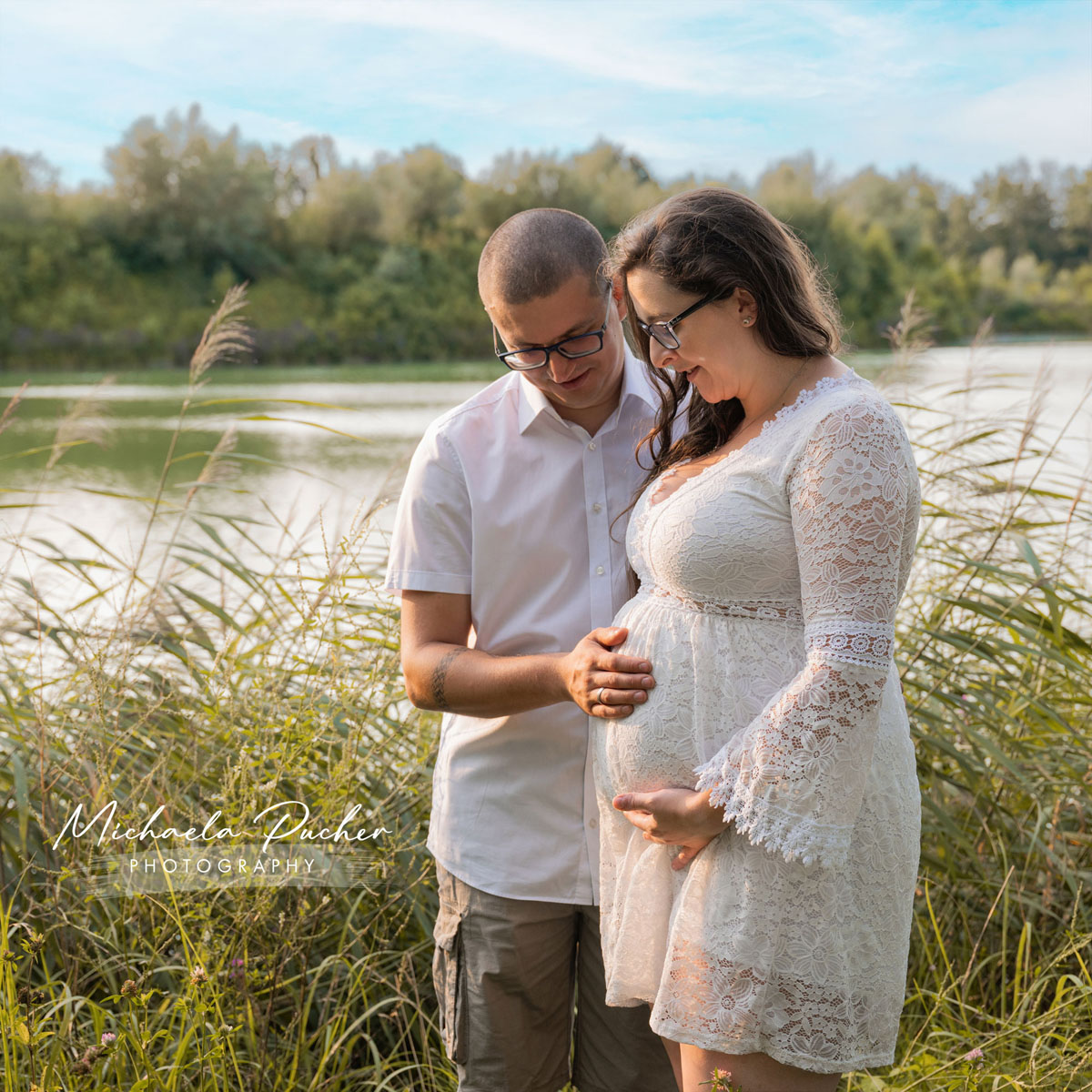 Paar steht am Seeufer, beide berühren liebevoll den Babybauch der werdenden Mama, umgeben von hohen Gräsern und warmem Abendlicht.